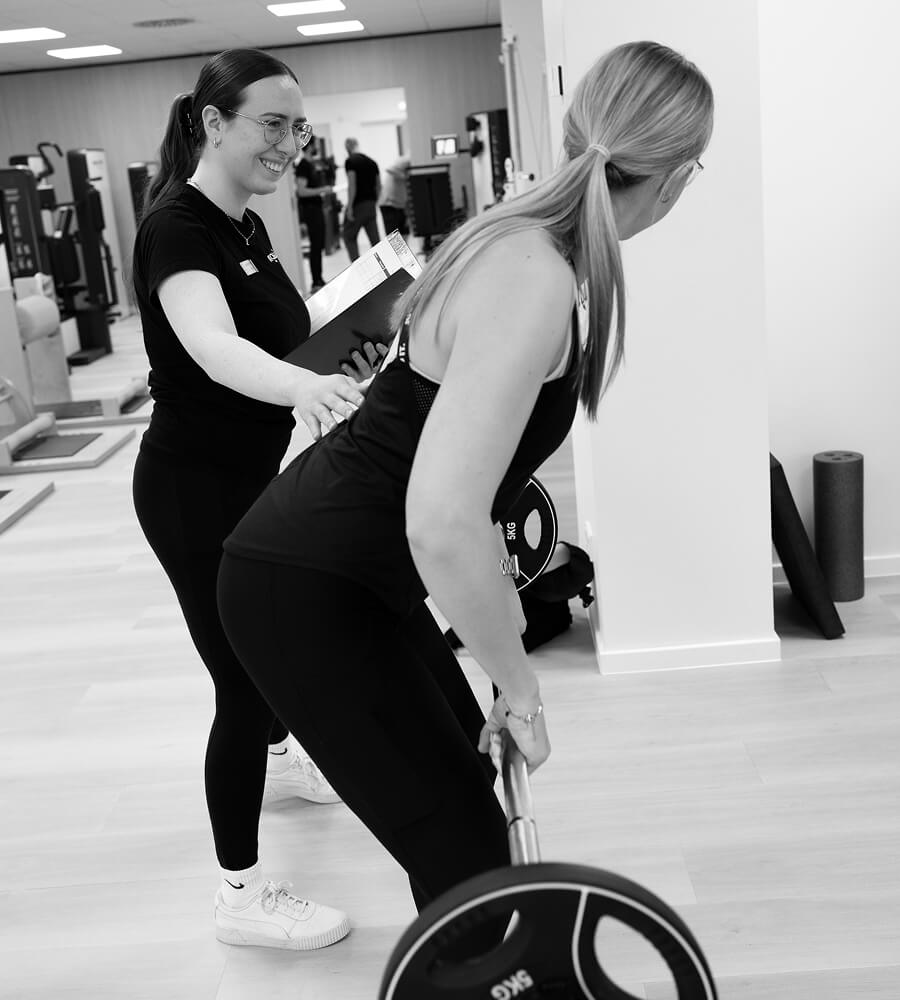 Frau hebt Langhantel unter Anleitung einer Trainerin – praktisches Training als Teil eines Praktikums in der Physiotherapie.